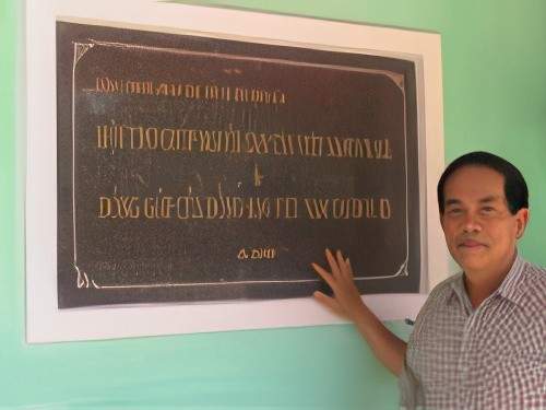 A man stands beside a large engraved plaque on a wall, touching it with his right hand. The plaque has text written in Vietnamese.