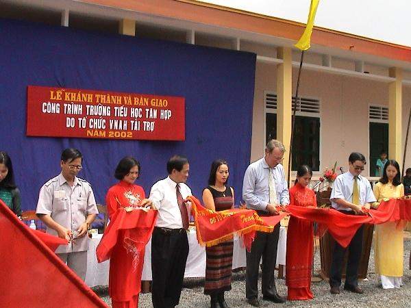 A group of people stand in a line, cutting a large red ribbon at an outdoor event in front of a blue and red banner.
