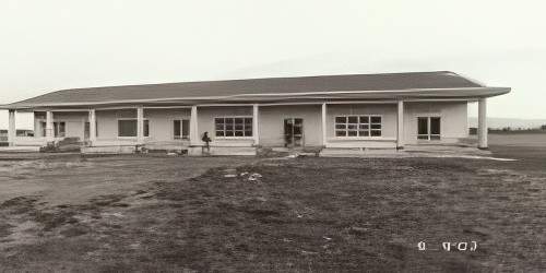 A sepia-toned photograph of a single-story building with a gable roof and multiple columns at the front. The building is surrounded by open, barren land.