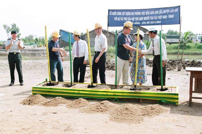 A group of people wearing construction hats are participating in a groundbreaking ceremony, standing on a platform and holding shovels. A banner with text is displayed in the background.