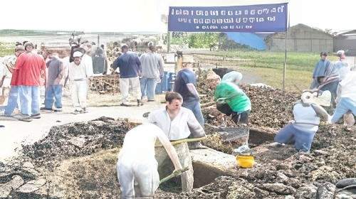 A group of people, some holding shovels, work on digging a large hole at an outdoor site with a blue sign in the background.