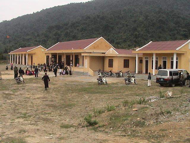 A group of people gather around several yellow buildings with red roofs in a rural, mountainous area.