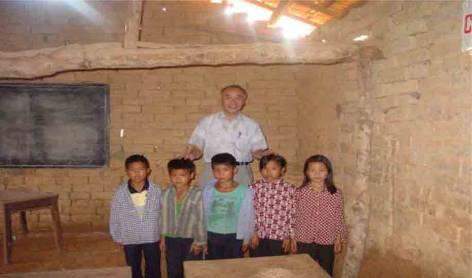 A teacher stands behind five children in a sparsely furnished classroom with earthen walls and a wooden ceiling.