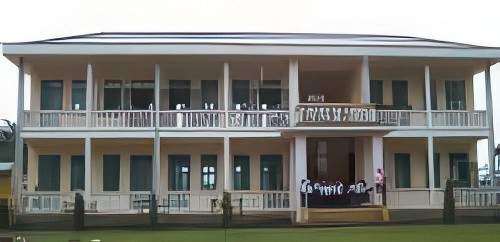 Two-story building with a balcony and multiple windows on both floors. A group of people stands on the steps at the entrance. The building is surrounded by grass.