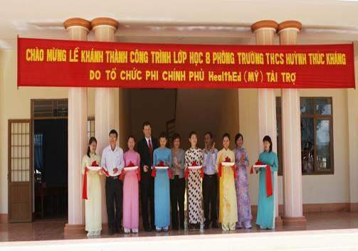 A group of people standing in front of a building under a red banner with Vietnamese text. They are dressed in formal attire and are holding a ribbon, suggesting a ceremonial event.