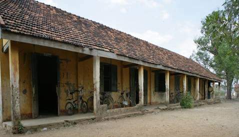 An old, single-story building with a clay-tiled roof and several stone pillars. The exterior shows signs of wear, and there is sparse vegetation around it.