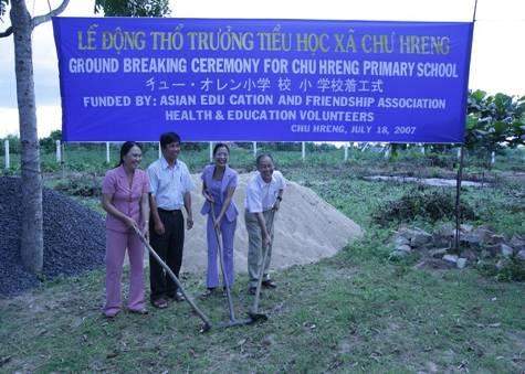 Four people stand with shovels at a groundbreaking ceremony for Chi Pheng Primary School, as indicated by a large blue sign behind them. They are standing on a site with piles of dirt.