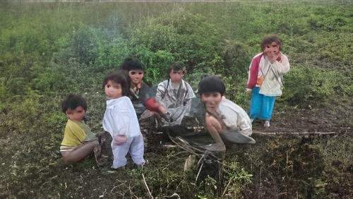 Six children are sitting and standing in a grassy field, some looking at the camera, with expressions of curiosity and contentment. The field is green with patches of dry soil.