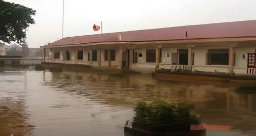 A single-story building with a red roof stands next to a flooded area. The building has multiple windows and is adorned with a flagpole displaying a red flag.