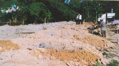 A construction site with workers and some equipment. Dirt and materials are visible on the ground with trees in the background.