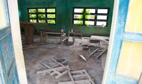An abandoned classroom with broken furniture and debris scattered on the floor, viewed through an open door. The windows are intact but dirty.