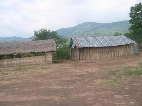 Two thatched-roof buildings stand in a rural area with a backdrop of hills and trees. The ground is bare with patches of grass.