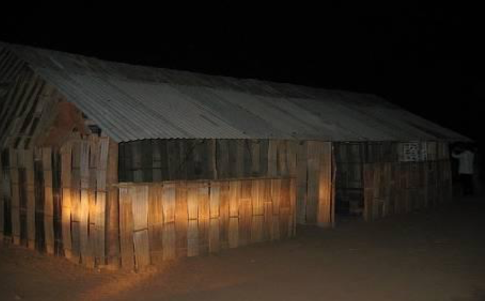 A dimly lit, simple wooden structure with a corrugated metal roof and walls made of vertical planks stands in the dark.
