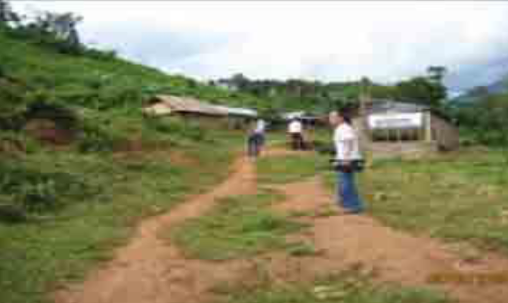 Three people walk on a dirt path towards several simple buildings in a rural, hilly area with lush greenery.