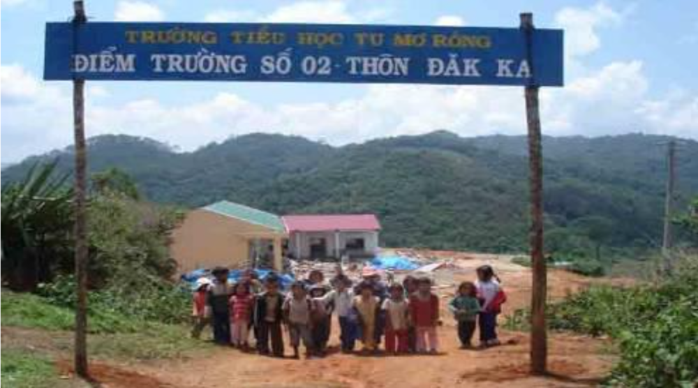 Group of children stand under a sign in Vietnamese at an outdoor location with mountains in the background. There are small buildings and piles of materials behind them.