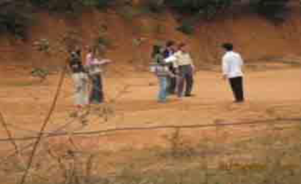 A group of people standing and conversing outdoors on a dirt path with shrubbery in the background.