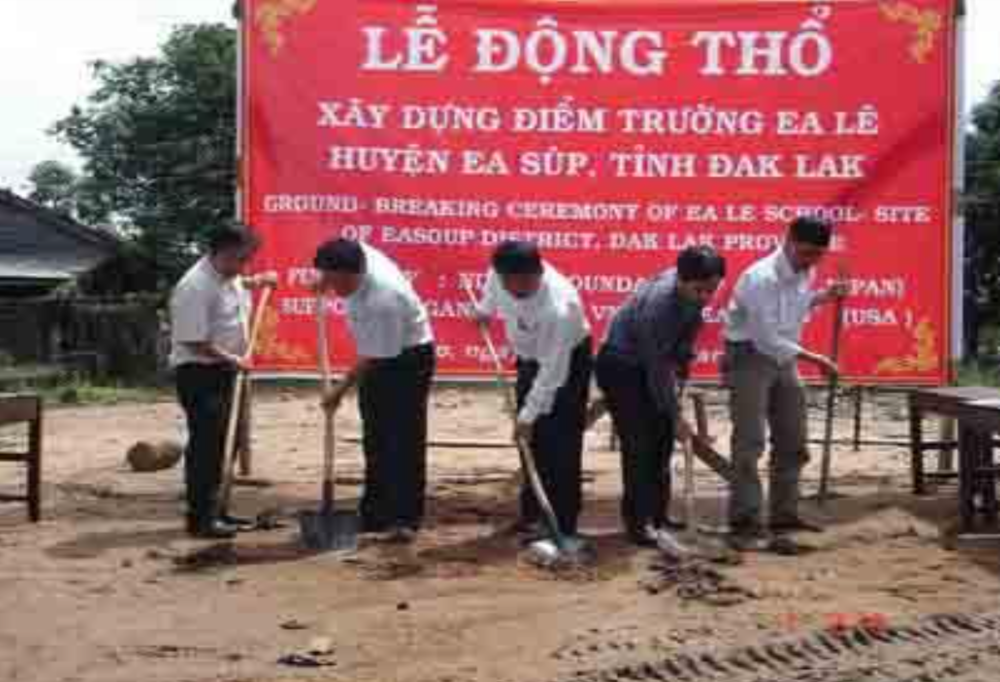 Five people, each holding a shovel, participate in a ground-breaking ceremony. They stand in front of a red banner with Vietnamese text about the construction of a new school in Ea Le, Dak Lak.