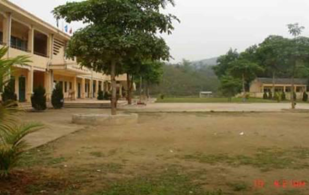 A school courtyard with a two-story building on the left, trees, and a clear sky. The ground is mostly dirt with some sparse grass patches.