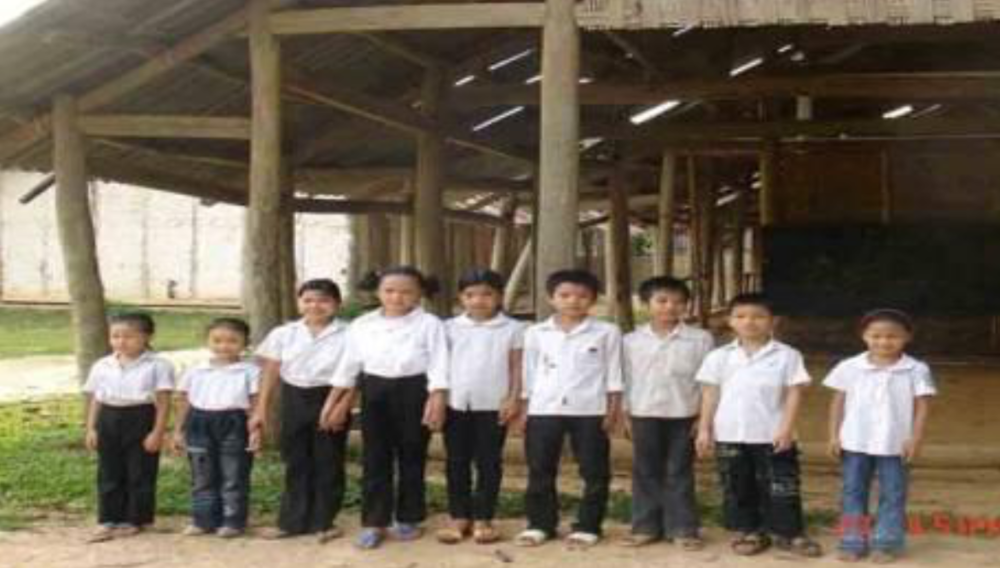 A group of ten children, dressed in similar white shirts and dark pants, stands in a line outside a wooden structure with a thatched roof.