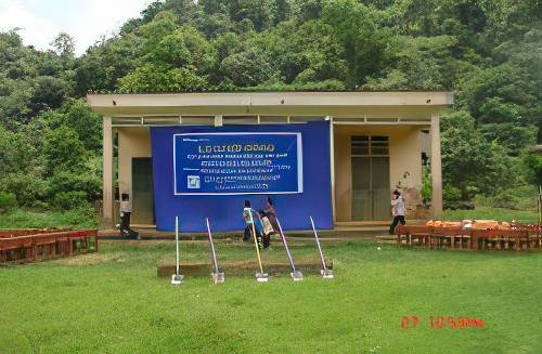 A small building with a large blue banner in front, situated in a grassy area with trees in the background. There are chairs and tools, including shovels, arranged in front of the building.