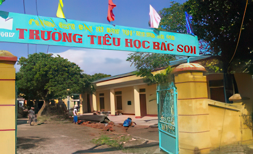 Entrance of Truong Tieu Hoc Bac Son with a few children playing outside. The blue gate is open, and colorful flags are flying above the entrance. Trees and a yellow building are visible inside.