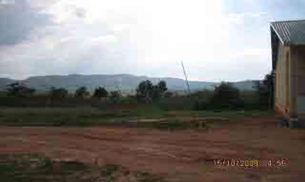 A dirt field with sparse vegetation, distant mountains, and a building partially visible on the right. The sky is mostly clear with some clouds. Time and date are displayed in the bottom right.