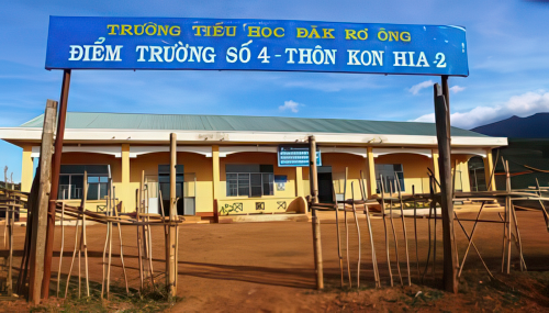 A yellow school building with a blue sign in Vietnamese, reading "TrÆ°á»ng Tiá»ƒu Há»c ÄÄƒk RÆ¡ Ã”ng, Äiá»ƒm TrÆ°á»ng Sá»‘ 4 - ThÃ´n Kon Hia 2," stands behind a fence on a dirt yard under a clear sky.