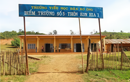 A small, single-story school building with a blue sign above the entrance. The sign has Vietnamese text and there is a wooden fence in the foreground. The area appears to be rural.