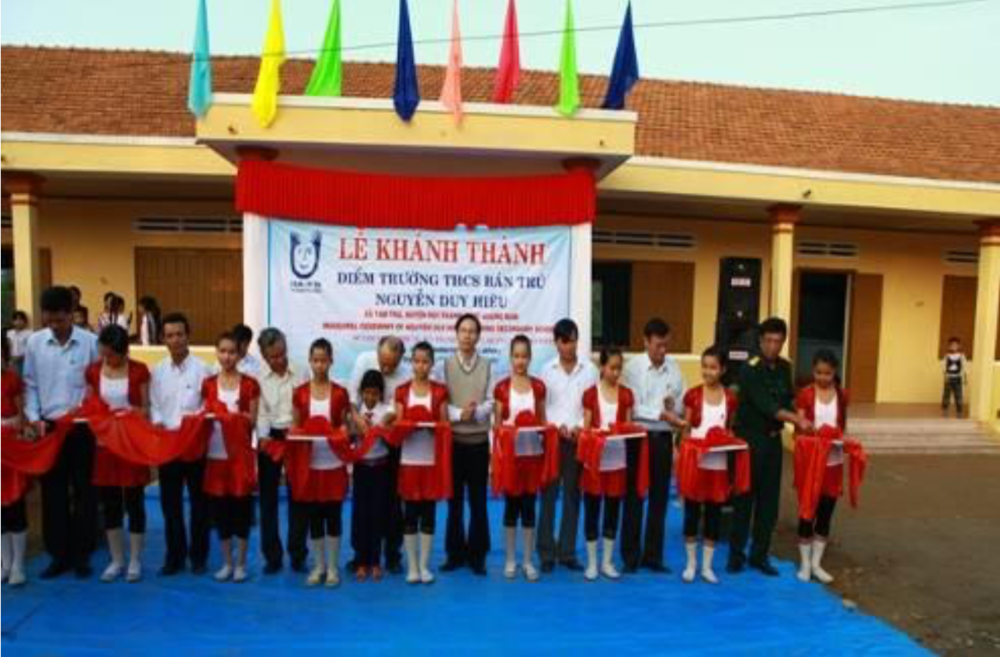 A group of people, including children in school uniforms, stand in a line holding red sashes at an event in front of a building. A banner is displayed behind them with Vietnamese text.
