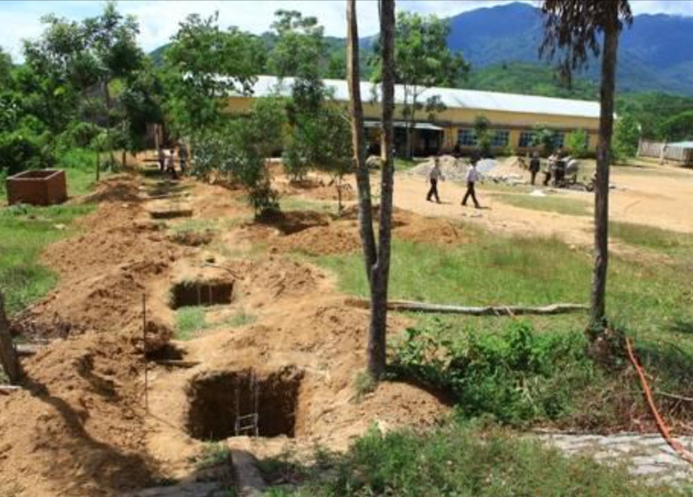 A construction site with multiple dug holes in the ground, surrounded by grassy areas. In the background, people walk near a yellow building with mountains visible in the distance.