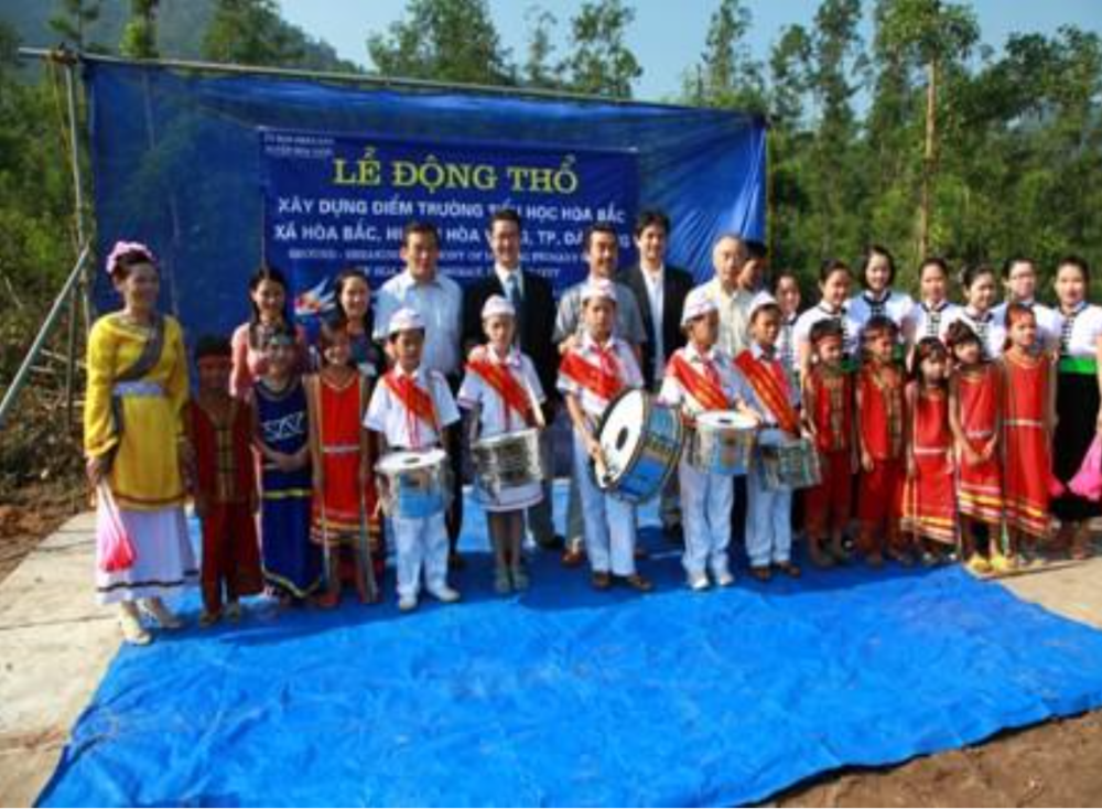 A group of children dressed in traditional attire hold musical instruments, posing with adults in business attire in front of a blue banner and a backdrop of trees.