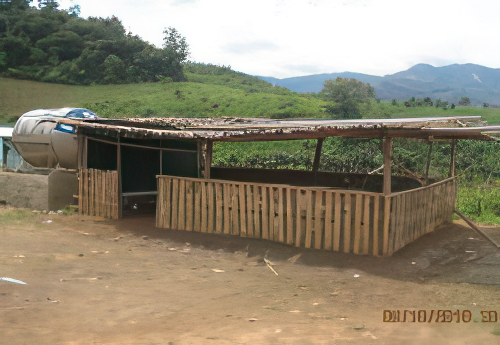 A small wooden shelter with an open side, a slanted roof, and a large water container beside it, set in a rural landscape with greenery and hills in the background.