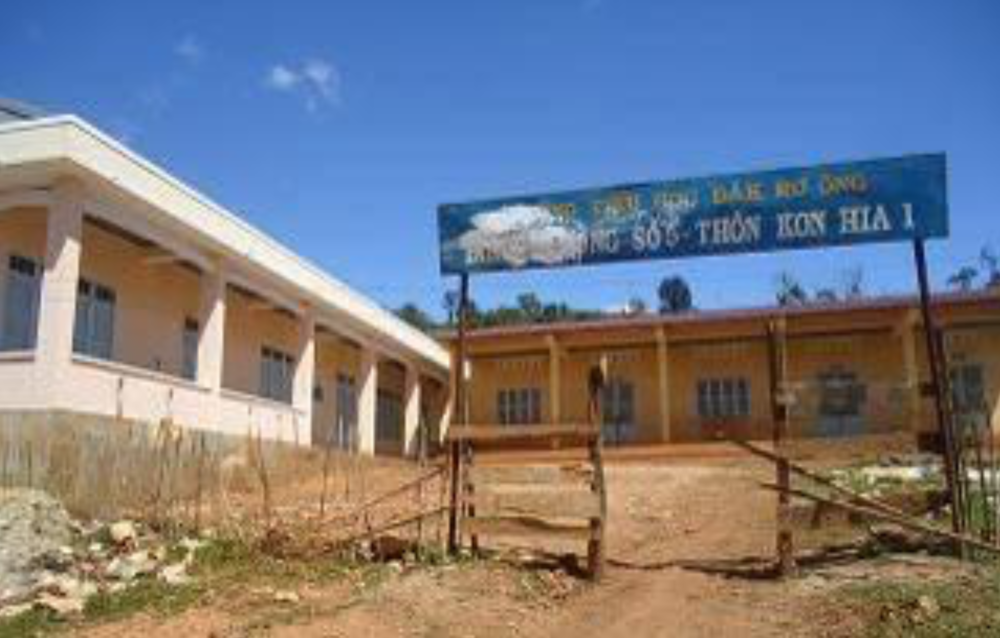 A dirt path leads to a school with a blue sign in Vietnamese at the entrance. Adjacent buildings have a simple, rectangular design. Clear sky overhead.