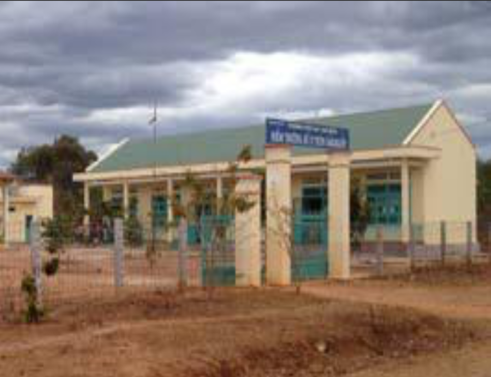 A school building with a sign above the entrance reading "TRUONG TIEU HOC LIEN SON DIEM TRUONG SO 2 THON DAKRIJOP." The area around the school appears rural with cloudy skies in the background.