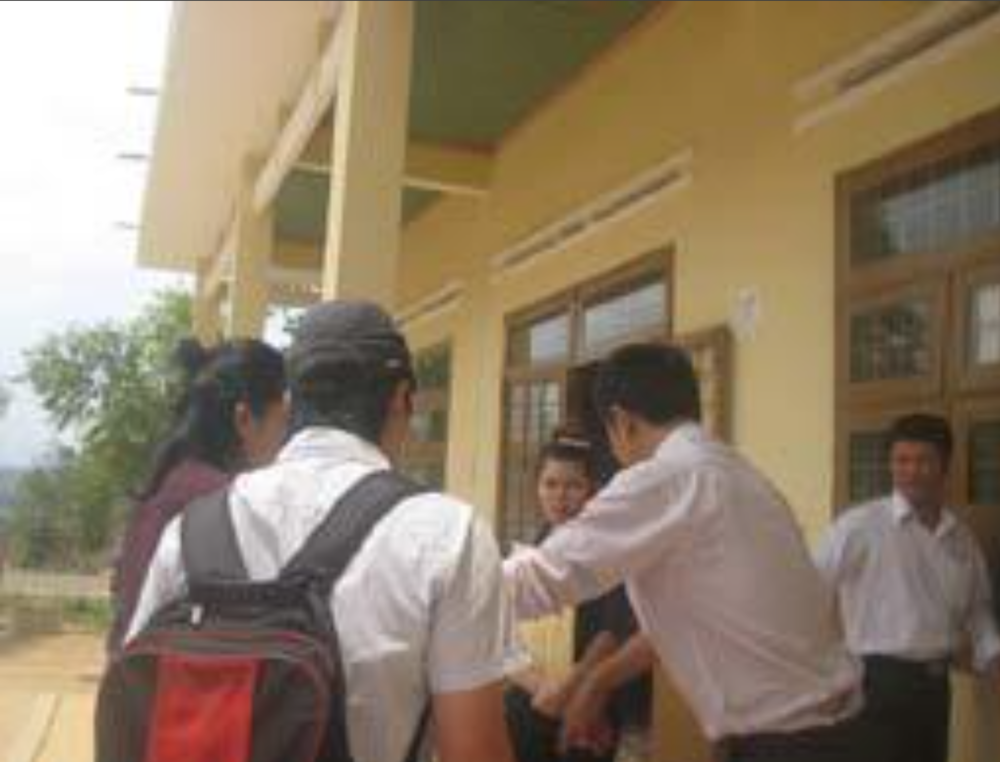 A group of people stand outside a yellow building. One person wears a backpack. Two individuals shake hands while others observe. Trees are visible in the background.