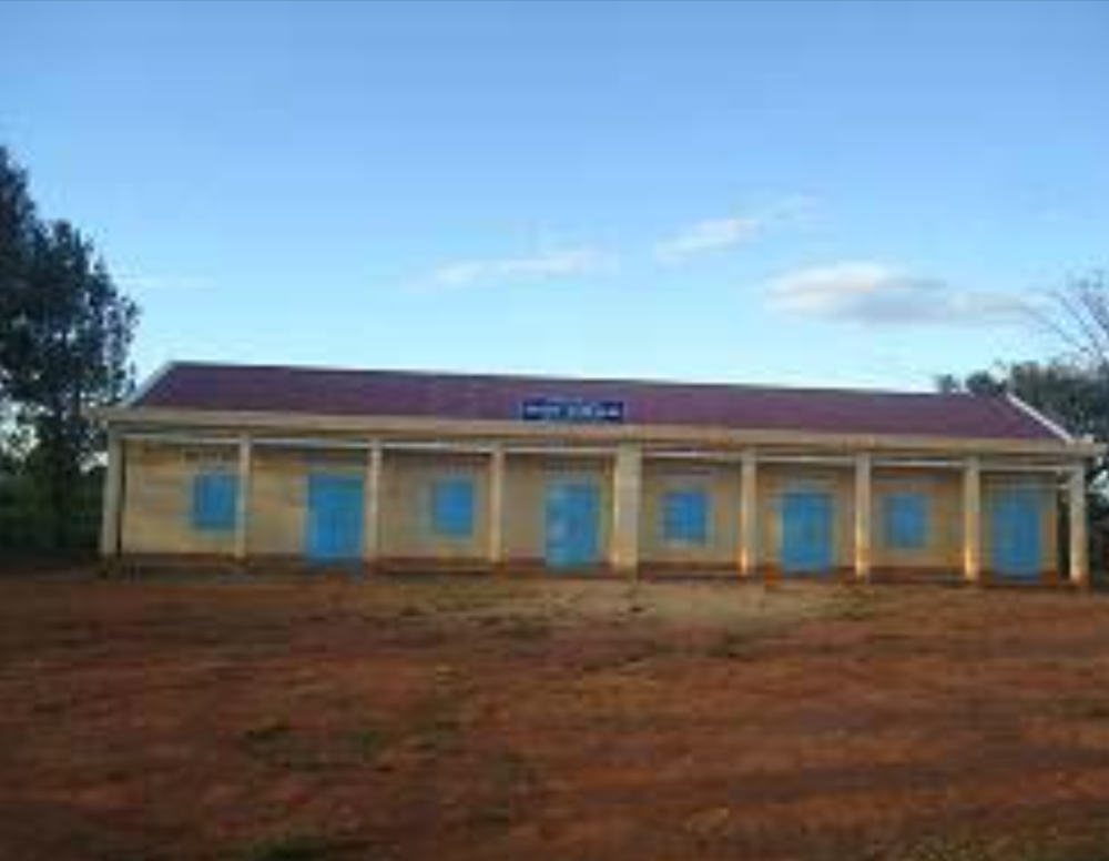 A single-story building with a red roof and light-colored walls, featuring several blue doors and windows. It is set on a brown dirt ground with trees visible on one side.