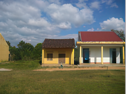Two small houses, one with a rusted brown roof and the other with a red roof, sit side by side on a grassy area under a partly cloudy sky.