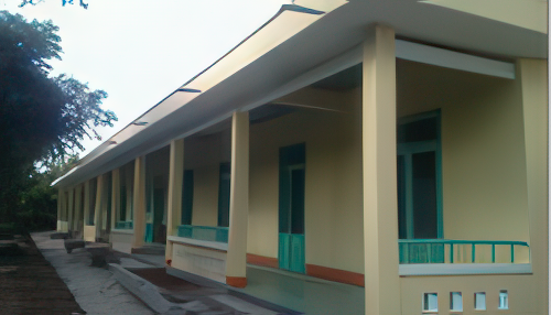 A single-story building with a long corridor and multiple green doors and windows, supported by pillars. The area around the building appears under-construction.