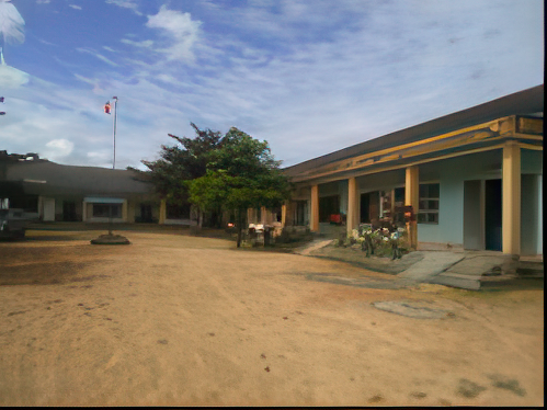 A school building with a sandy courtyard, surrounded by trees under a partly cloudy sky. A flagpole with a flag is visible in the background.