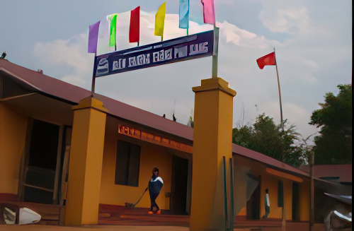 A school building with a sign and colorful flags above the entrance. Two people are standing near the doorway, and a flagpole with a red flag is in the background. Trees are visible on the right.