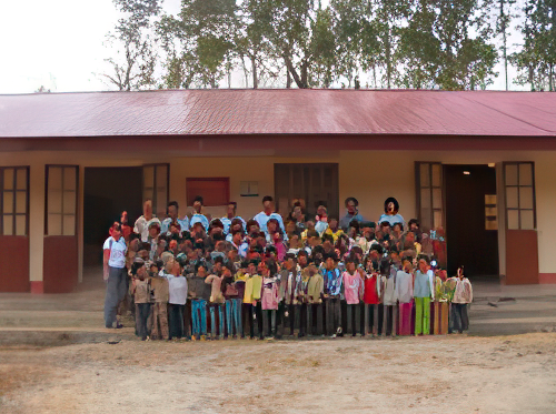 A large group of children and adults stand in front of a building with a red roof and several doors, posing for a photo. Trees are visible in the background.