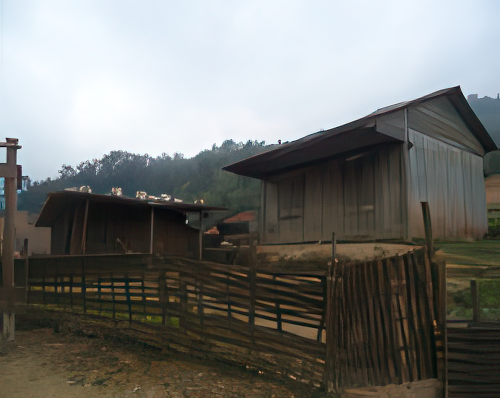 Two wooden sheds with tin roofs stand in front of a hillside. A rustic wooden fence encloses a small area in front of the structures. Overcast sky above.