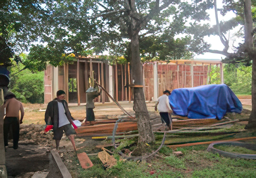 Construction workers building a structure in a wooded area, with some workers carrying wooden planks and others working near a partially built frame. A blue tarp covers a pile of materials nearby.