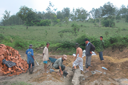 A group of people work on a construction site in a rural area, with brick piles and tools visible. They are surrounded by greenery and trees in the background.