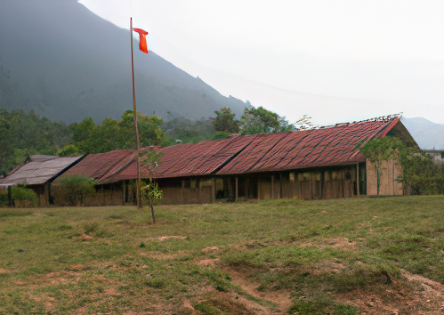 A row of abandoned, weathered buildings with rusted metal roofs and a small orange flag on a pole in front. They are set in a grassy area with trees and a mountain in the background.