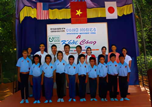 A group of people, including adults and children in uniform, stand together on a stage adorned with flags from multiple countries, with a sign featuring various symbols and text in the background.