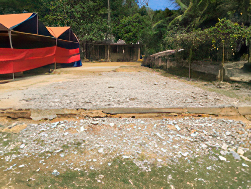 A partially damaged concrete pavement with debris scattered around. Colorful tents are visible to the left, surrounded by green vegetation and trees.