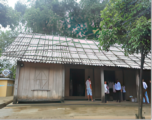 A group of people stand outside a rustic, wooden house with a sloped roof in a forested area.